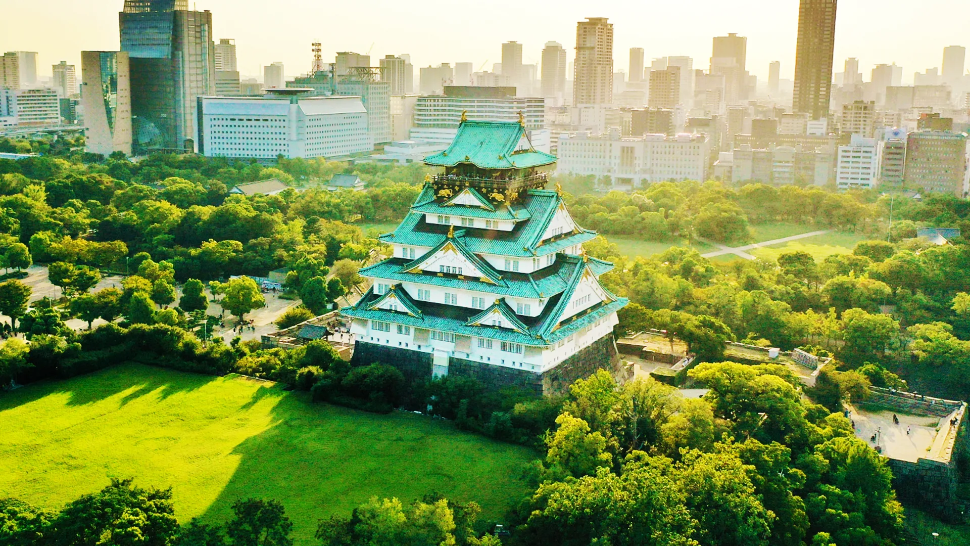 Aerial view of Osaka Castle's main keep in its park, with the city skyline behind it